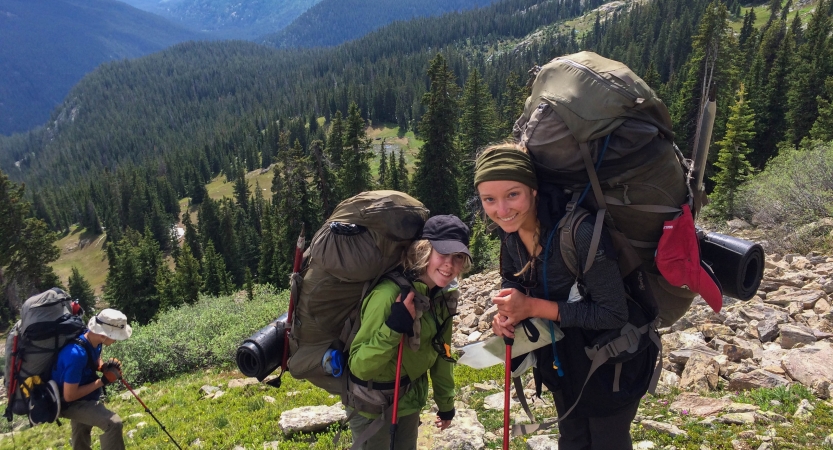 three people with backpacks make their way up a rocky and grassy incline